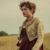 "Woman in historical dress standing in a field with tall grass, trees in the background, and a cloudy sky, creating a calm, pastoral setting."