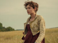 "Woman in historical dress standing in a field with tall grass, trees in the background, and a cloudy sky, creating a calm, pastoral setting."