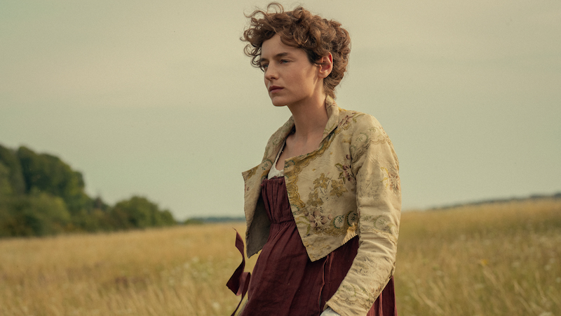 "Woman in historical dress standing in a field with tall grass, trees in the background, and a cloudy sky, creating a calm, pastoral setting."