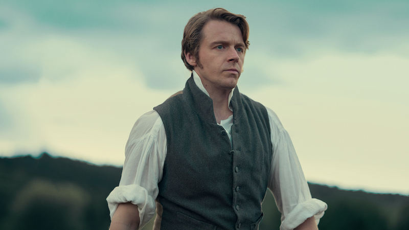 "A man in historical clothing stands outdoors with a serious expression, wearing a vest over a white shirt, with hills and trees in the blurred background under a cloudy sky."