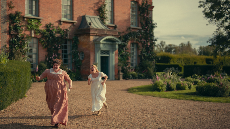 "Two women in period dresses run joyfully on a gravel path in front of a large brick house with ivy and flowers, surrounded by hedges and gardens under a partly cloudy sky."