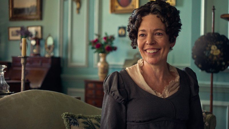 "A smiling woman in period costume sits on a couch in a vintage, elegant living room with teal walls, antique furniture, framed art, and a flower vase in the background."